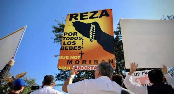 Protesters holding signs. One sign has hands holding rosary.