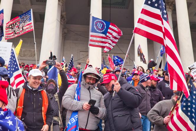 Men holding different flags such as Trump flags and 3 percenters flag. Two men in the front are looking at a phone