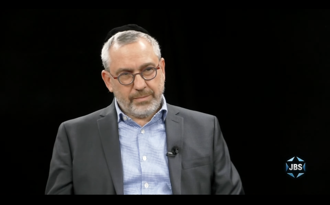 White man with glasses sitting against a black background