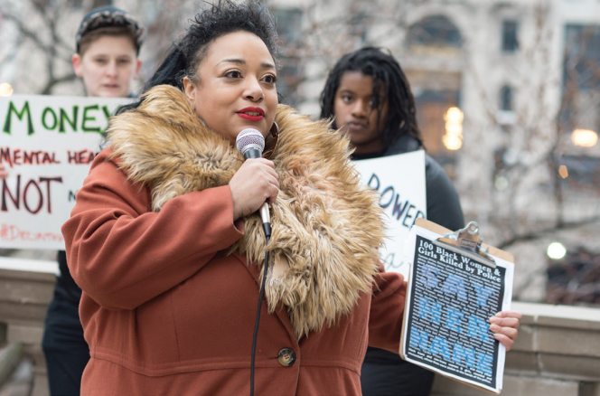 A person speaking with a Say Her Name sign