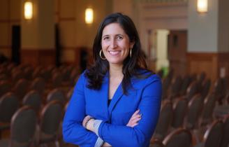 Smiling woman with long brown hair, gold hoop earrings, and a blue blazer, arms crossed