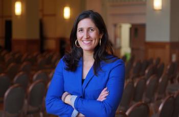 Smiling woman with long brown hair, gold hoop earrings, and a blue blazer, arms crossed