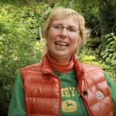 A White woman with short blond hair wearing a green t-shirt and orange sleeveless jacket sitting in front of some bushes.