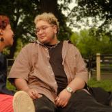 Two trans people in a park, looking at each other, laughing.