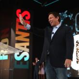 White man on stage with hand raised smiling, you can see the letters "ESANTIS" projected behind him.