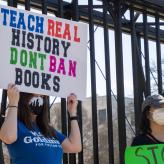 Woman in blue shirt holding a sign that reads "Teach real history don't ban books"