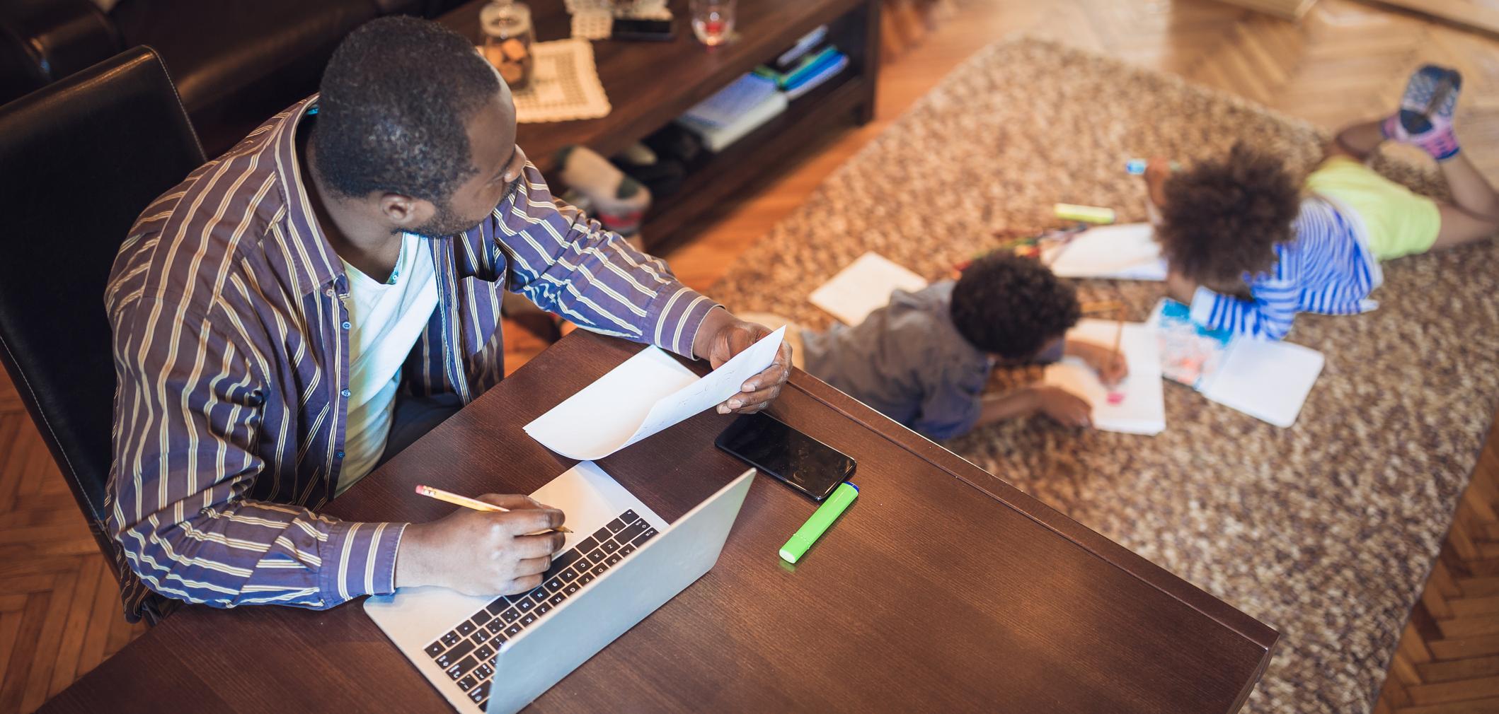 Adult man works on laptop and looks after two children reading nearby in room.