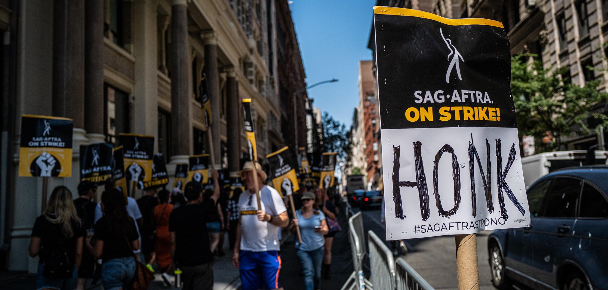 Protesters in the background. The Foreground has a sign that says "SAF-Aftra On Strike! Honk"