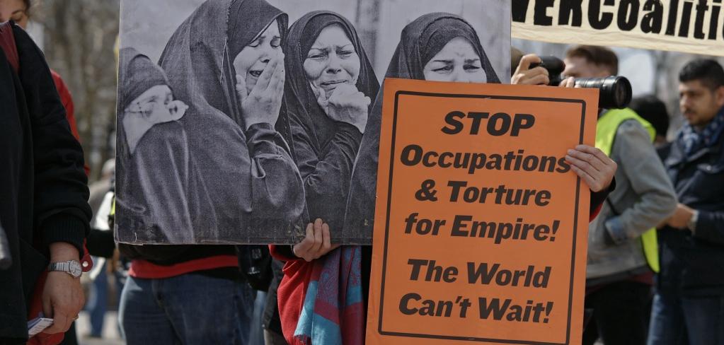 A person holding an image of women crying and a sign that says "stop occupations and torture for empire! The world can't wait!"