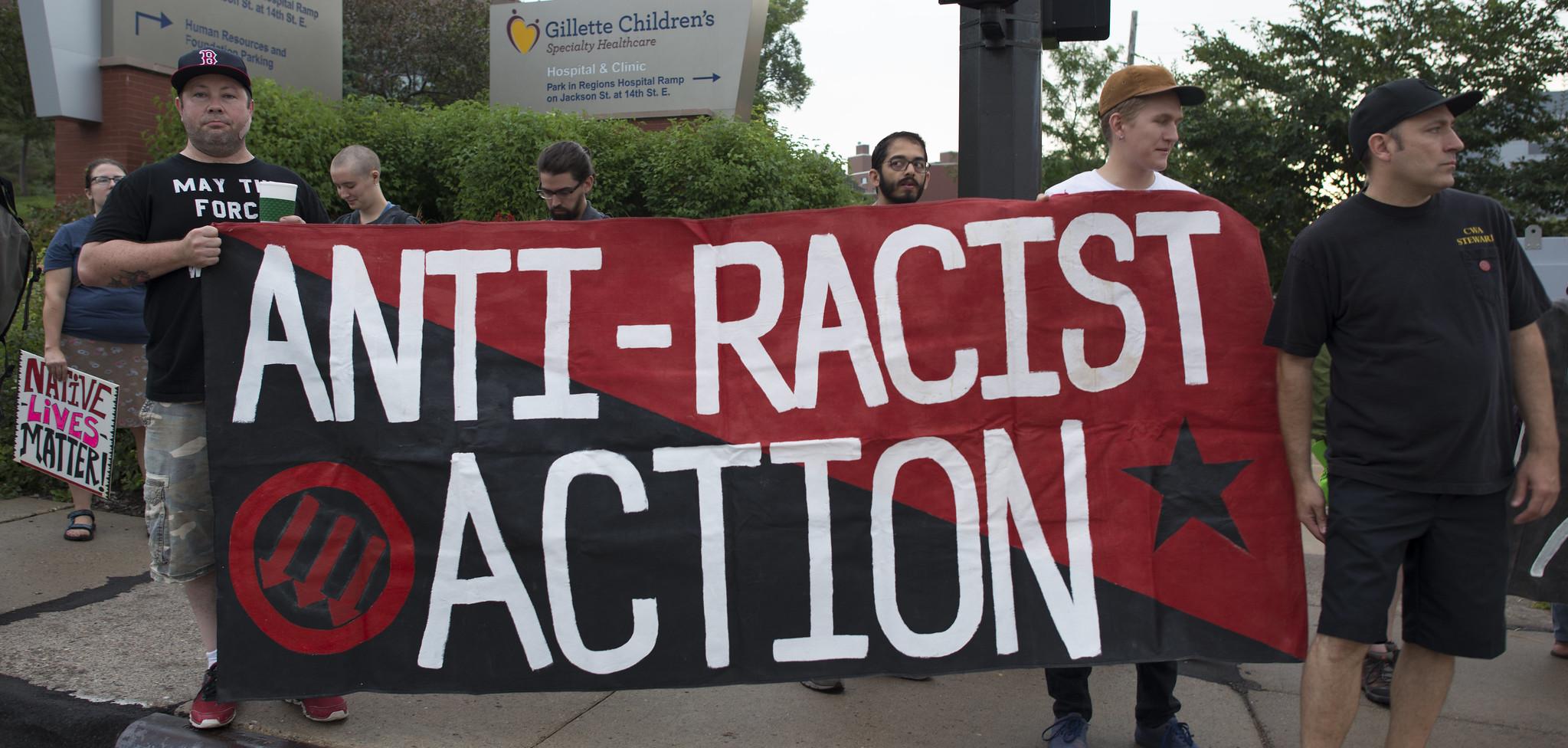 People holding a red and black flag that reads Anti-Racist Action