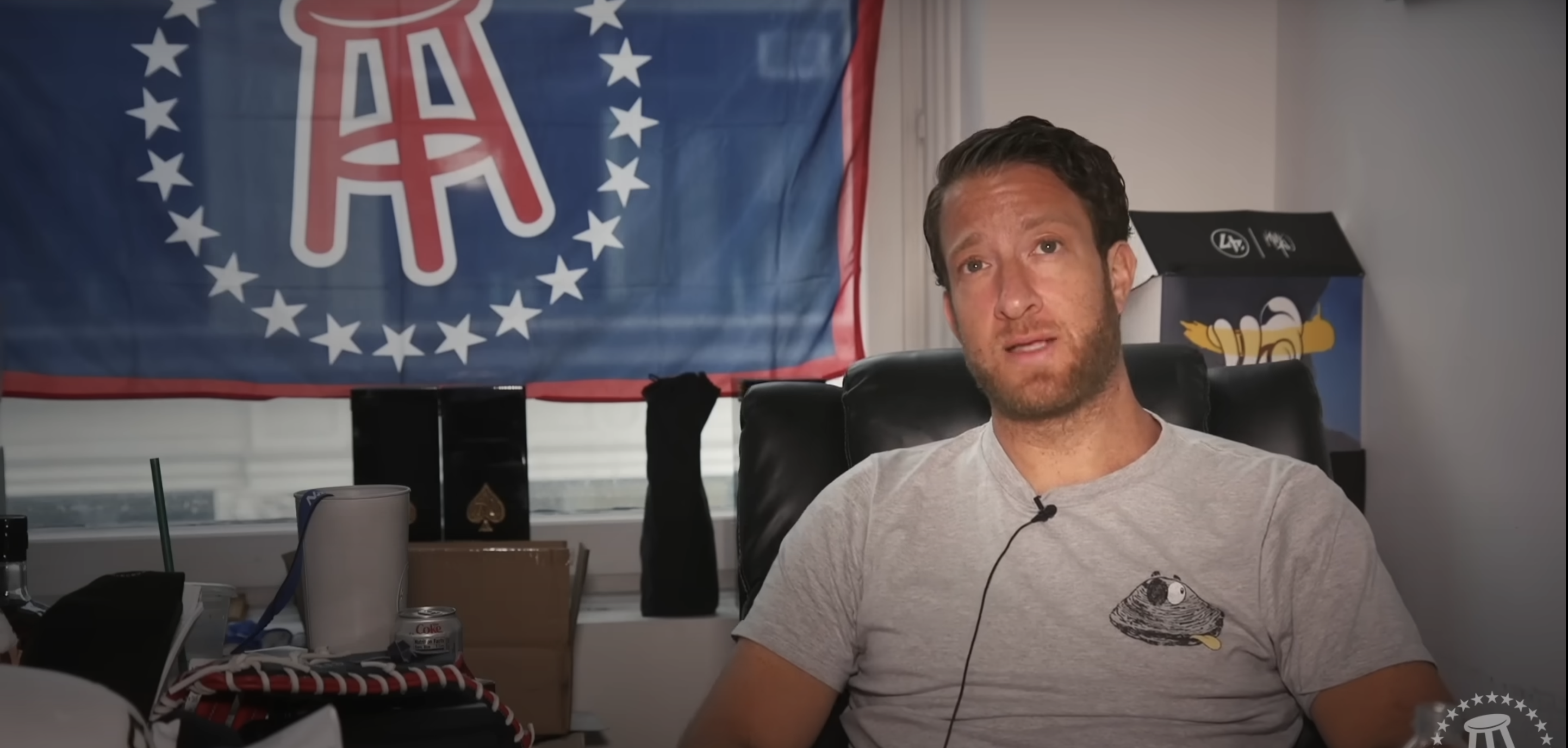 A man in a grey T-shirt sitting in a chair with a flag behind him with the Barstool logo