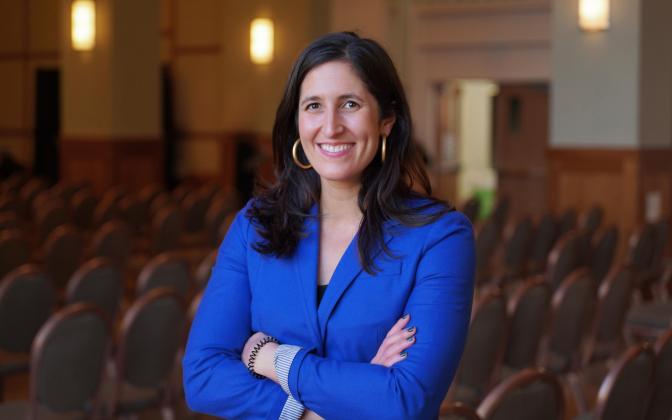 Smiling woman with brown hair, gold hoop earrings, a bright blue blazer, with arms crossed